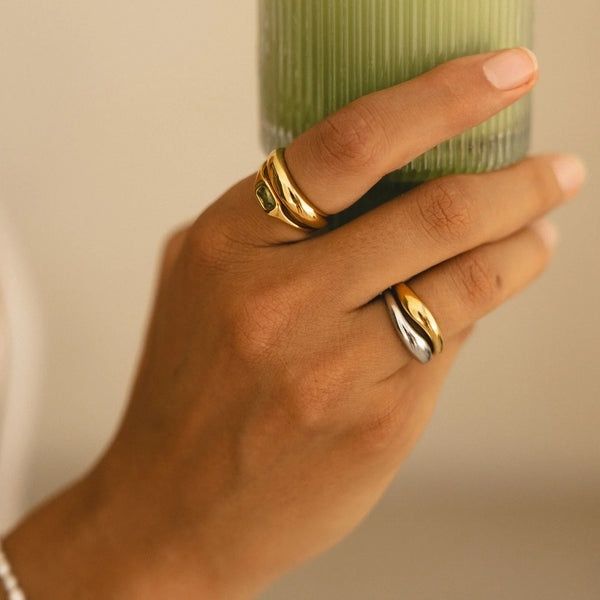 Hand med staplade ringar i guld och silver framför ett grönt glas